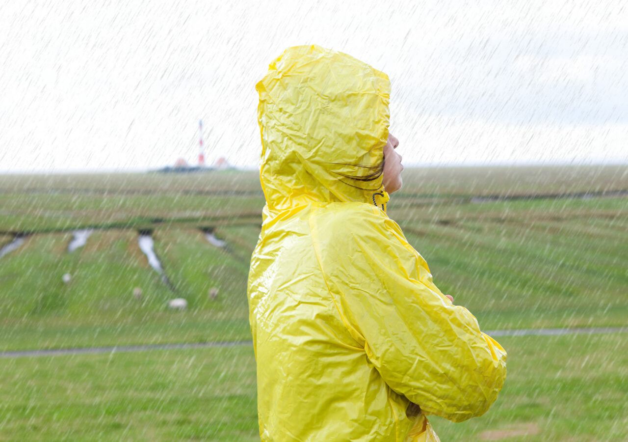 Eine Frau steht sichtlöich frustriert im Regen. Im Hintergrund ist der unscharf der Westerhever Leuchtturm zu sehen.